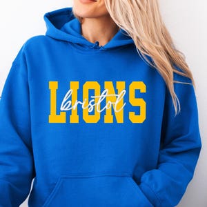 May include: A royal blue hoodie with a front pocket and a drawstring hood. The word "LIONS" is printed in yellow block letters, with "bristol" in a cursive font underneath. The person is wearing light blue jeans.