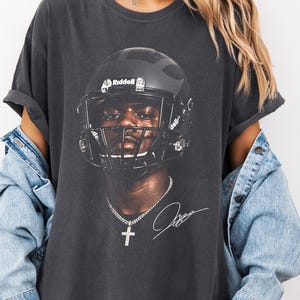 May include: Dark gray t-shirt featuring a football player wearing a black helmet. The image includes a signature and a cross pendant. The shirt is worn with a light blue denim jacket.