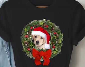 May include: Black t-shirt with a Christmas wreath design featuring a golden retriever puppy wearing a Santa hat. The wreath is decorated with red berries and green pine branches.