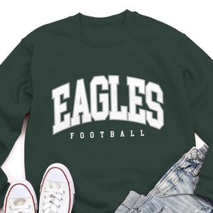 May include: A dark green sweatshirt with the word "EAGLES" in white with a white outline, and the word "FOOTBALL" in white underneath. The sweatshirt is laid flat on a white background with a pair of white trainers and a pair of light blue jeans.