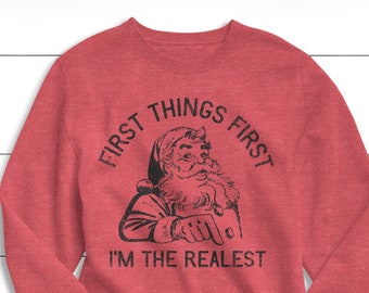 May include: Red sweatshirt with a black graphic of Santa Claus pointing. The text above the image reads "FIRST THINGS FIRST" and below the image, "I'M THE REALEST". The long-sleeved shirt is made of a soft material.