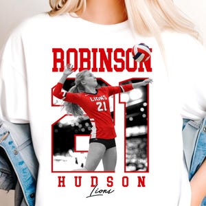 May include: White t-shirt featuring a volleyball player in a red uniform with the number 21. The name "ROBINSON" is printed above the image in red block letters, with "HUDSON Lions" below. The volleyball player is in action, spiking the ball.