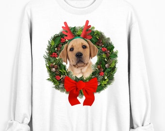May include: White sweatshirt featuring a Labrador Retriever puppy wearing a reindeer antler headband and framed by a Christmas wreath. A large red bow is at the bottom of the wreath. The sweatshirt is hanging on a wooden hanger.