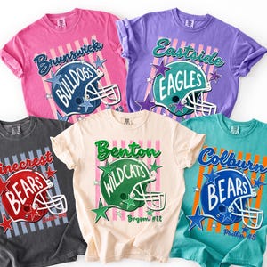 K&ouml;nnte beinhalten: F&uuml;nf kurz&auml;rmelige T-Shirts in Pink, Lila, Anthrazit, Creme und T&uuml;rkis. Jedes zeigt eine Football-Helm-Grafik, vertikale Streifen, Sternakzente und Teamnamen wie Brunswick Bulldogs, Eastside Eagles, Pinecrest Bears, Benton Wildcats und Colburn Bears.