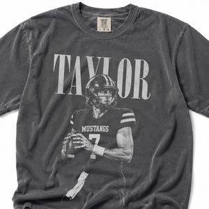 May include: A charcoal grey short-sleeve t-shirt featuring a graphic print of a football player wearing a helmet and a jersey with the text MUSTANGS 7. The name TAYLOR is printed in large white letters above the player. The shirt has a vintage wash.