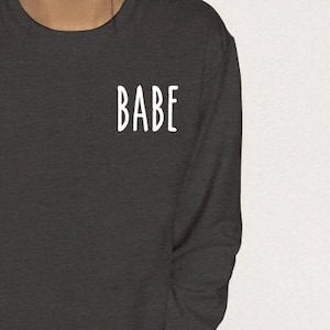 May include: A dark gray sweatshirt with long sleeves and a crew neck. The word "BABE" is printed in white on the front of the sweatshirt.