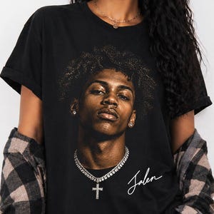 May include: Black t-shirt featuring a portrait of a person with curly hair, a silver chain, and a cross pendant. The name "Jalen" is written in cursive below the portrait. The person is wearing a black and brown plaid shirt.
