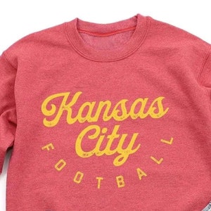 K&ouml;nnte beinhalten: Roter Pullover mit einer gelben Grafik, auf der "Kansas City Football" in einer Retro-Schrift steht.