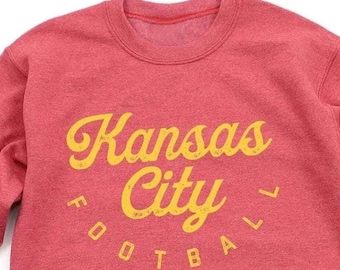 Puede incluir: Sudadera roja con un gráfico amarillo que dice "Kansas City Football" en una fuente retro.