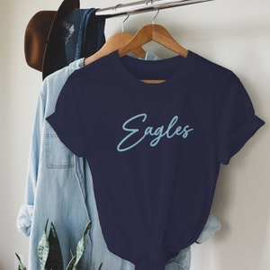 May include: A navy blue t-shirt with a light blue cursive inscription that reads "Eagles".