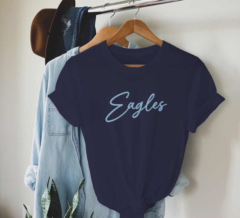 May include: A navy blue t-shirt with a light blue cursive inscription that reads "Eagles".