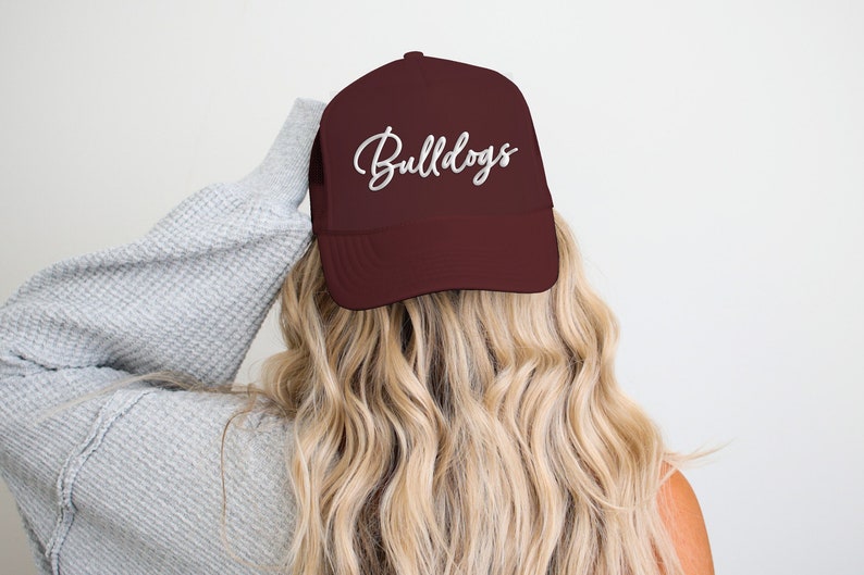 May include: A maroon baseball cap with the word "Bulldogs" in white script. The cap is worn by a person with long, wavy blonde hair and a gray sweater.