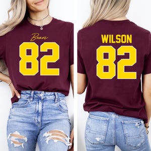 May include: Maroon t-shirt with the word "Bears" and the number "82" in yellow on the front. The back of the shirt displays the name "WILSON" and the number "82" in yellow. The shirt is paired with blue jeans.