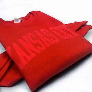 May include: Red sweatshirt with the text "KANSAS CITY" in a bold, raised font.