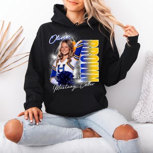 May include: Black hooded sweatshirt featuring a photo of a cheerleader with the name "Olivia Brown" and "Mustang Cheer" printed on it. The design includes blue and yellow accents.