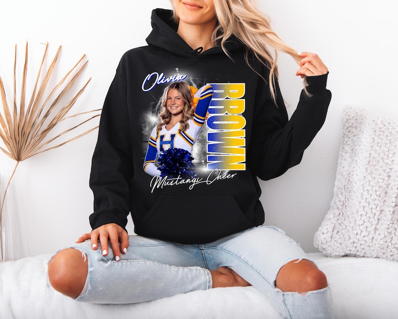 May include: Black hooded sweatshirt featuring a photo of a cheerleader with the name "Olivia Brown" and "Mustang Cheer" printed on it. The design includes blue and yellow accents.