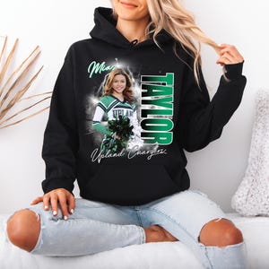 May include: Black hoodie featuring a cheerleader graphic. The design includes the name "TAYLOR" in green and white, along with the words "Upland Chargers" and a photo of a cheerleader. The hoodie has a drawstring hood and a front pocket.