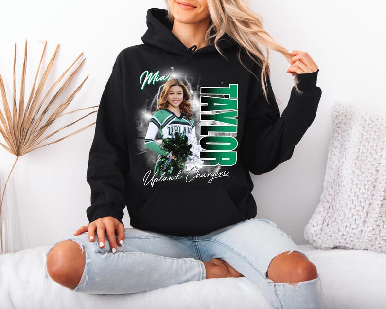 May include: Black hoodie featuring a cheerleader graphic. The design includes the name "TAYLOR" in green and white, along with the words "Upland Chargers" and a photo of a cheerleader. The hoodie has a drawstring hood and a front pocket.