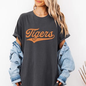 May include: Dark gray t-shirt with the word "Tigers" in orange script. The shirt has a vintage, slightly distressed look. The text is arched and has a subtle shadow effect. The shirt is a casual, oversized style.