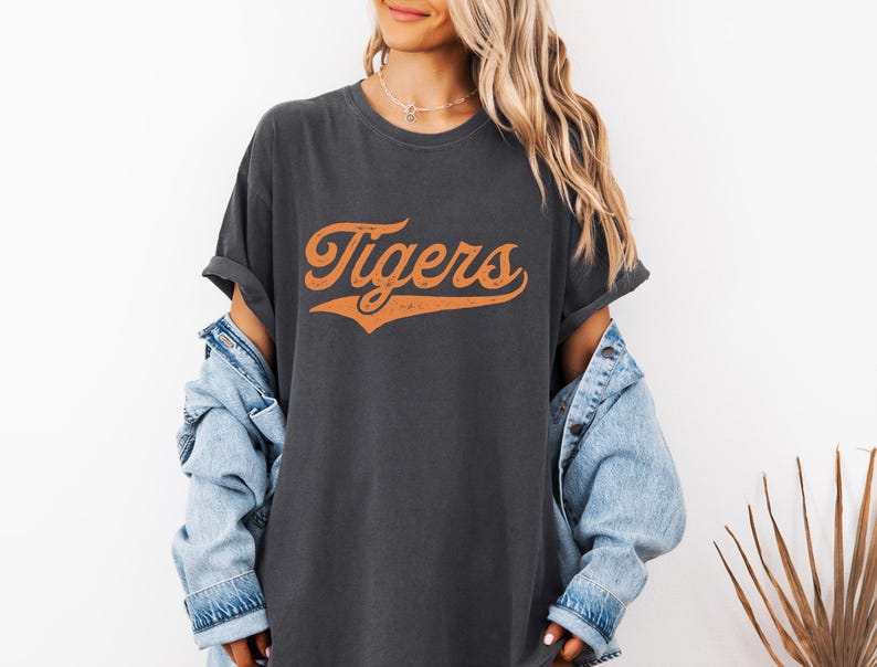 May include: Dark gray t-shirt with the word "Tigers" in orange script. The shirt has a vintage, slightly distressed look. The text is arched and has a subtle shadow effect. The shirt is a casual, oversized style.