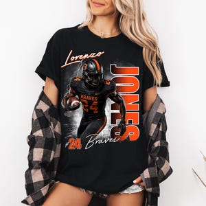 K&ouml;nnte beinhalten: Ein schwarzes T-Shirt mit einer Grafik eines Footballspielers in schwarz-orangem Trikot mit der Nummer 24. Der Text lautet Lorenzo, Jones und #24 Braves. Das Design zeigt einen Football, Blitze und einen rauchigen Hintergrundeffekt.