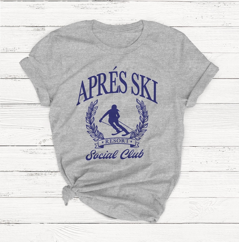 May include: Gray t-shirt with a blue graphic of a skier in a wreath with the text "Apr&egrave;s Ski Resort Social Club".