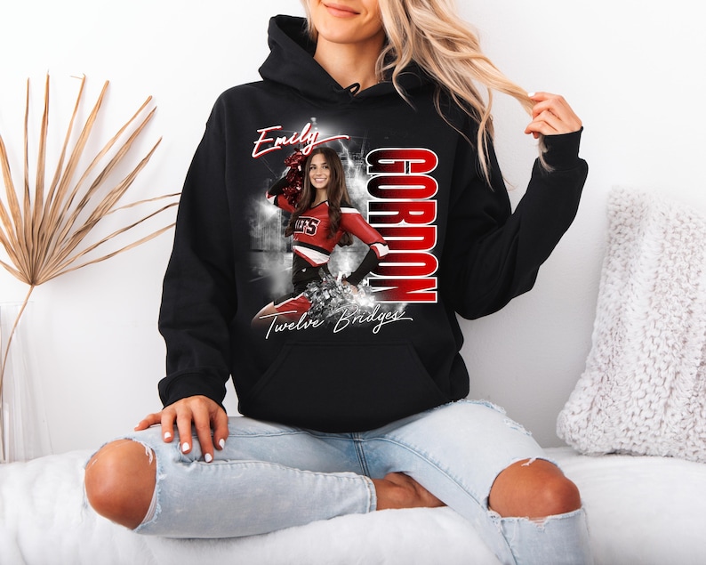 May include: Black hooded sweatshirt featuring a graphic of a cheerleader in a red and white uniform. The sweatshirt has the name "Emily" in white script, the name "Gordon" in red block letters, and "Twelve Bridges" in script.
