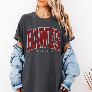 May include: A charcoal gray short-sleeve t-shirt featuring the word HAWKS in large, bold, maroon collegiate-style lettering with a white outline, and the word FOOTBALL in smaller white text underneath, worn with a light blue denim jacket.