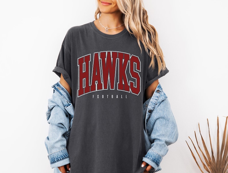 May include: A charcoal gray short-sleeve t-shirt featuring the word HAWKS in large, bold, maroon collegiate-style lettering with a white outline, and the word FOOTBALL in smaller white text underneath, worn with a light blue denim jacket.