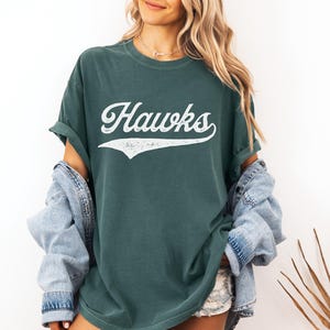 May include: Dark green t-shirt with the word "Hawks" in white script. The shirt has a vintage, worn look. The model is wearing a denim jacket over the shirt.