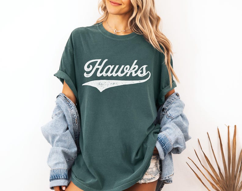 May include: Dark green t-shirt with the word "Hawks" in white script. The shirt has a vintage, worn look. The model is wearing a denim jacket over the shirt.