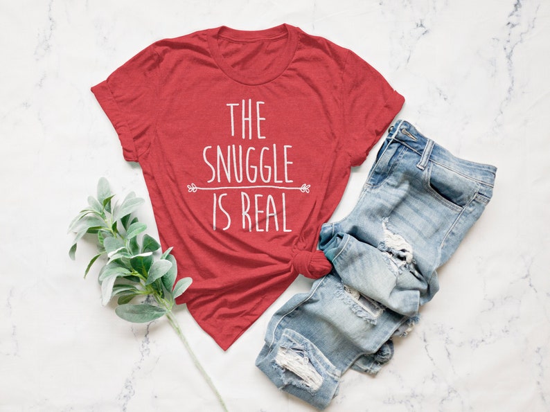 May include: Red t-shirt with white text that reads "The Snuggle is Real". The shirt is paired with blue jeans.