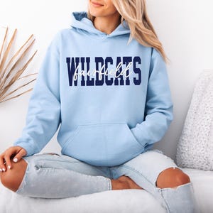 May include: A light blue hoodie with the word "WILDCATS" in navy blue block letters and "Fairfield" in white script. The hoodie has a front pocket and a hood. The model is wearing light blue jeans.