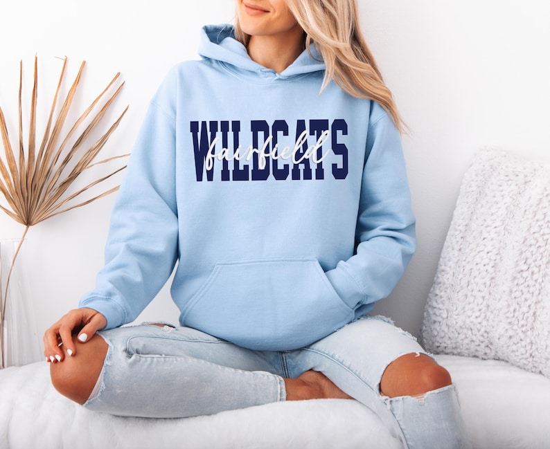May include: A light blue hoodie with the word "WILDCATS" in navy blue block letters and "Fairfield" in white script. The hoodie has a front pocket and a hood. The model is wearing light blue jeans.