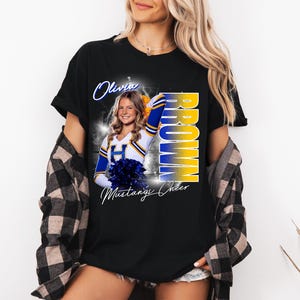 May include: Black t-shirt featuring a cheerleader in a blue and gold uniform. The shirt has the name "Olivia Brown" and "Mustangs Cheer" printed on it. The cheerleader is holding blue pom-poms.