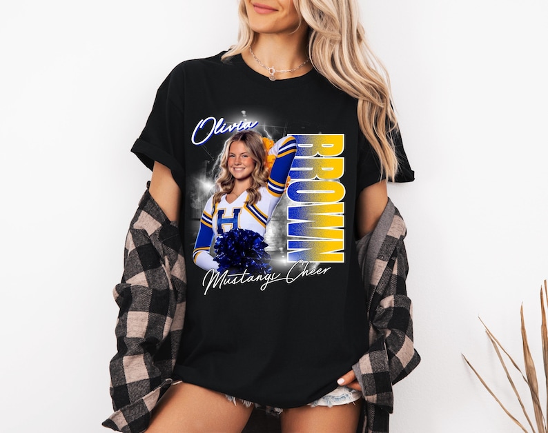 May include: Black t-shirt featuring a cheerleader in a blue and gold uniform. The shirt has the name "Olivia Brown" and "Mustangs Cheer" printed on it. The cheerleader is holding blue pom-poms.