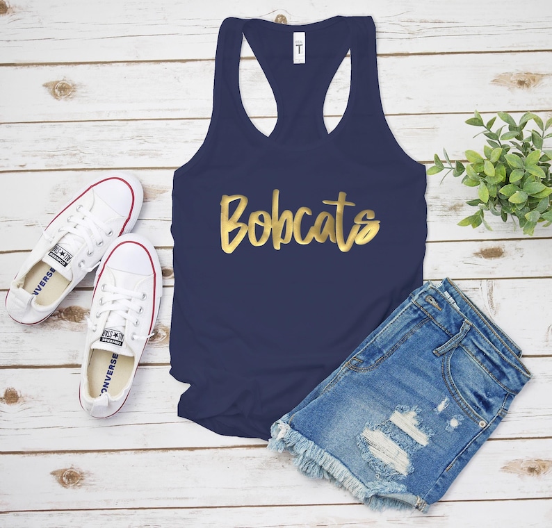 May include: A navy blue racerback vest with the gold text "Bobcats". The image also shows denim shorts and white Converse trainers with red trim. A small green plant is also visible.
