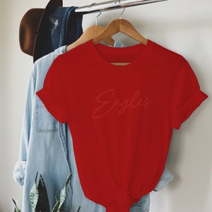 May include: Red short-sleeved t-shirt with the word "Eagles" written in a cursive font.