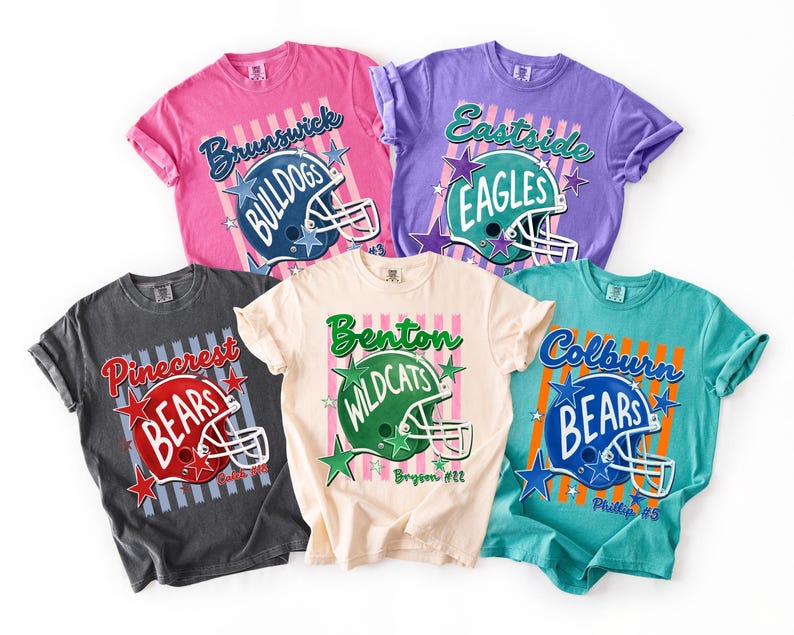 K&ouml;nnte beinhalten: F&uuml;nf kurz&auml;rmelige T-Shirts in Pink, Lila, Anthrazit, Creme und T&uuml;rkis. Jedes zeigt eine Football-Helm-Grafik, vertikale Streifen, Sternakzente und Teamnamen wie Brunswick Bulldogs, Eastside Eagles, Pinecrest Bears, Benton Wildcats und Colburn Bears.