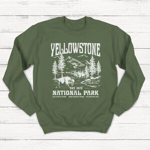 May include: Green sweatshirt with a white graphic design of Yellowstone National Park. The graphic includes a bison, trees, mountains, and a river. The text reads "Yellowstone National Park Est. 1872 Old Faithful Geyser Grand Prismatic Spring Yellowstone Lake."