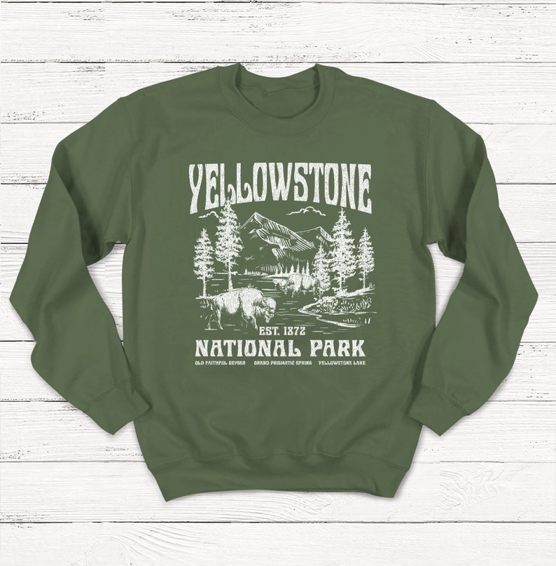 May include: Green sweatshirt with a white graphic design of Yellowstone National Park. The graphic includes a bison, trees, mountains, and a river. The text reads "Yellowstone National Park Est. 1872 Old Faithful Geyser Grand Prismatic Spring Yellowstone Lake."