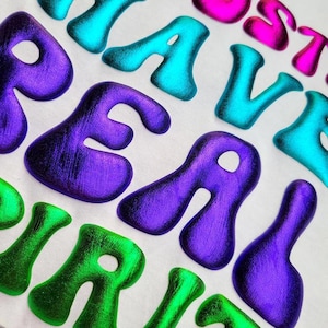 May include: Close-up of white fabric featuring the words "REAL VIBES" and "MOSTLY" in a retro, bubble letter font. The letters are in shades of purple, teal, green, and pink.