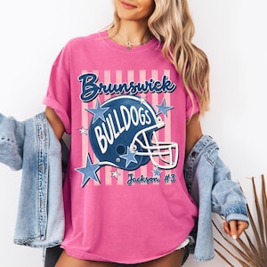 K&ouml;nnte beinhalten: Ein leuchtend pinkes T-Shirt mit einer Grafik eines dunkelblauen Football-Helms und dem Schriftzug Brunswick Bulldogs. Das Design enth&auml;lt hellrosa vertikale Streifen, blaue Sterne und den Text Jackson #3 in einer Schreibschrift auf der Vorderseite.