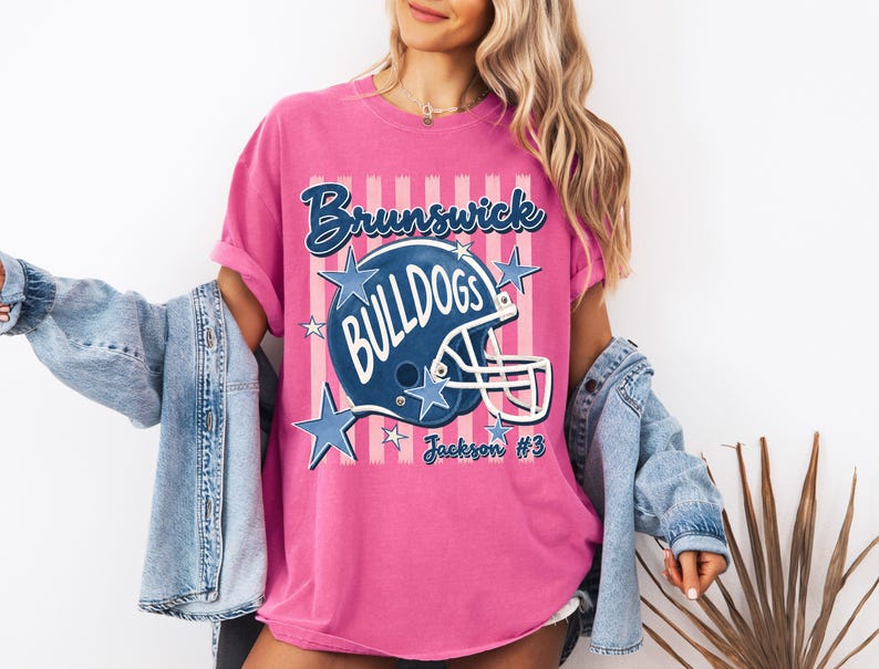 K&ouml;nnte beinhalten: Ein leuchtend pinkes T-Shirt mit einer Grafik eines dunkelblauen Football-Helms und dem Schriftzug Brunswick Bulldogs. Das Design enth&auml;lt hellrosa vertikale Streifen, blaue Sterne und den Text Jackson #3 in einer Schreibschrift auf der Vorderseite.