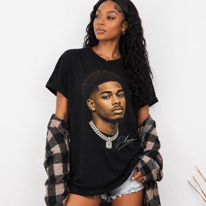 May include: Black t-shirt featuring a detailed portrait of a man with a chain necklace and a signature. The graphic is in shades of brown and tan. The shirt is worn with a plaid shirt and denim shorts.