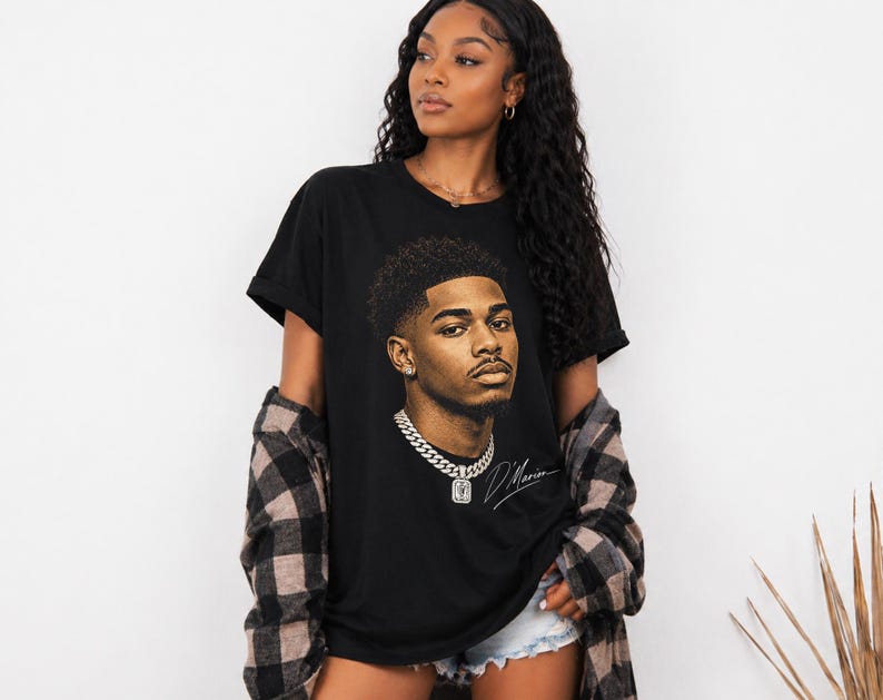 May include: Black t-shirt featuring a detailed portrait of a man with a chain necklace and a signature. The graphic is in shades of brown and tan. The shirt is worn with a plaid shirt and denim shorts.