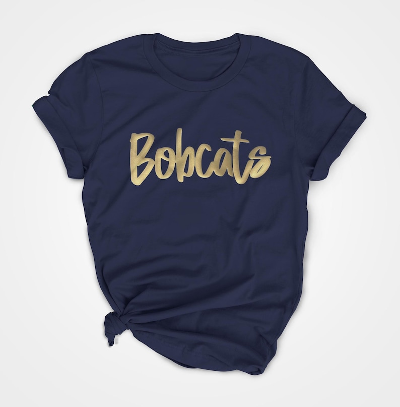 May include: A navy blue t-shirt featuring the word "Bobcats" in gold script. The t-shirt is folded and knotted at the bottom, with the sleeves rolled up.