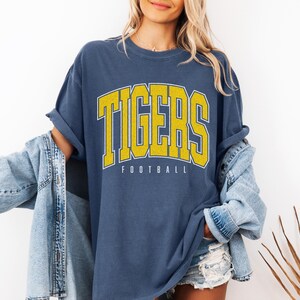 May include: A blue short-sleeved t-shirt featuring the word TIGERS in large yellow block letters with a white outline, positioned above the word FOOTBALL in smaller white text. The shirt is worn with a light blue denim jacket and denim shorts.