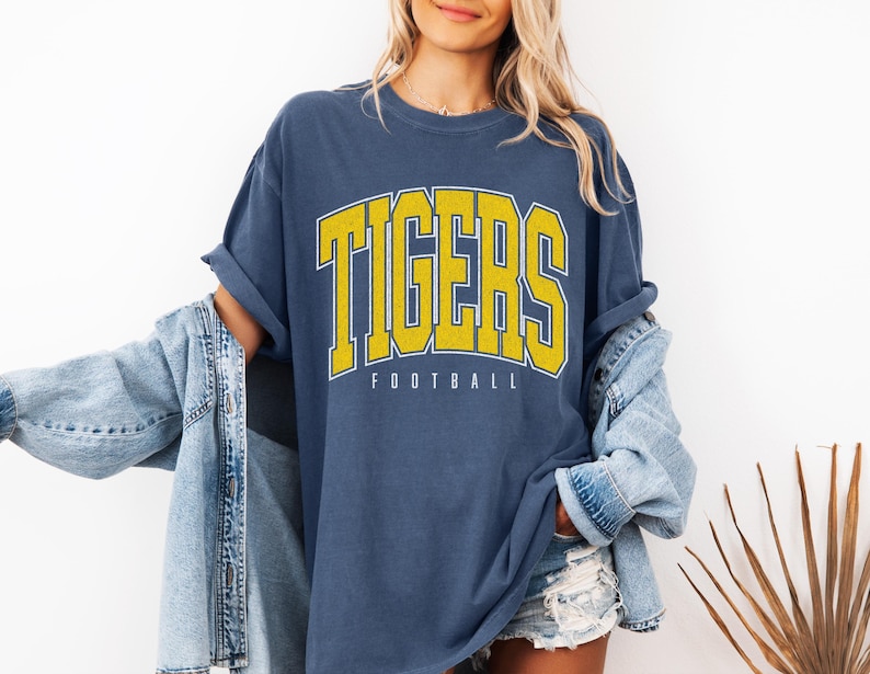 May include: A blue short-sleeved t-shirt featuring the word TIGERS in large yellow block letters with a white outline, positioned above the word FOOTBALL in smaller white text. The shirt is worn with a light blue denim jacket and denim shorts.