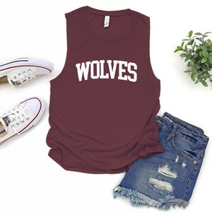 May include: A maroon sleeveless shirt with the word "WOLVES" in white block letters. The shirt is paired with denim shorts and white canvas sneakers. A small green plant is also in the image.